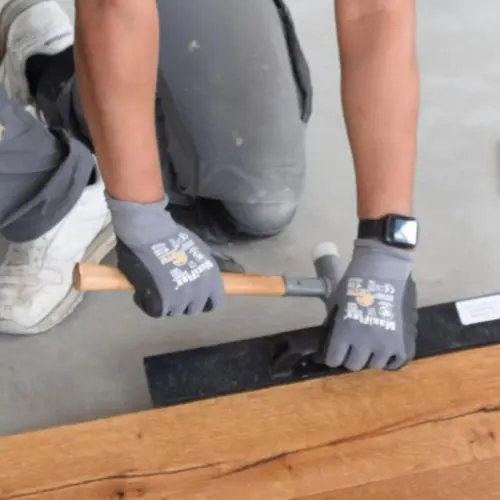 Handwerker mit Arbeitshandschuhen passt Holzboden mit Hammer und Schlagklotz auf einer glatten Betonfläche ein. Im Bild sind detailliert Werkzeuge und das strukturierte Holz sichtbar. Die Szene zeigt den präzisen Arbeitsablauf beim Verlegen von Bodenbelägen auf vorbereiteten Untergrund.